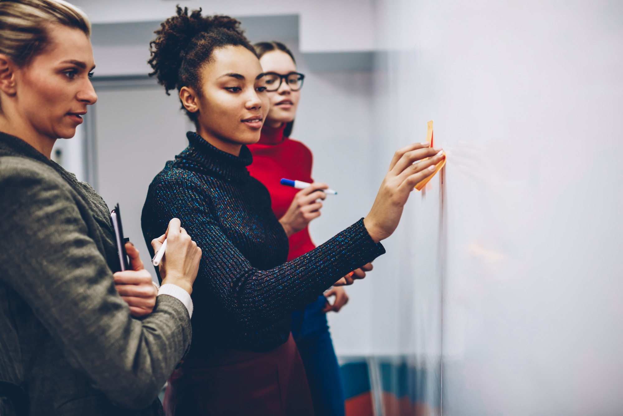 Three women collaborate at a whiteboard in an office setting. One writes with a marker while the others observe and discuss, conveying teamwork and focus.