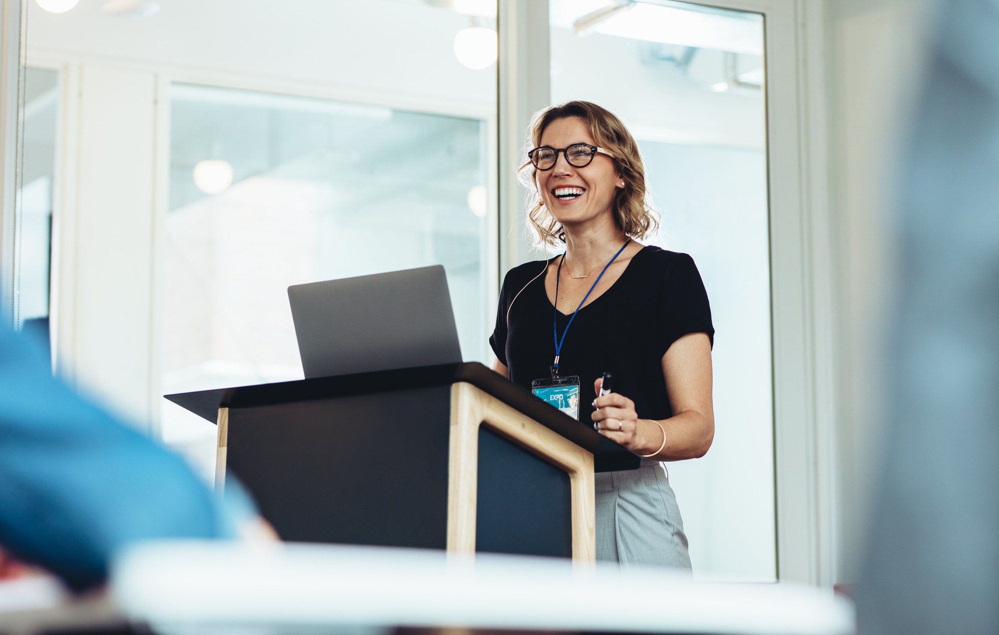 Smiling woman with glasses stands at a podium with a laptop, speaking confidently to an audience in a bright office setting.