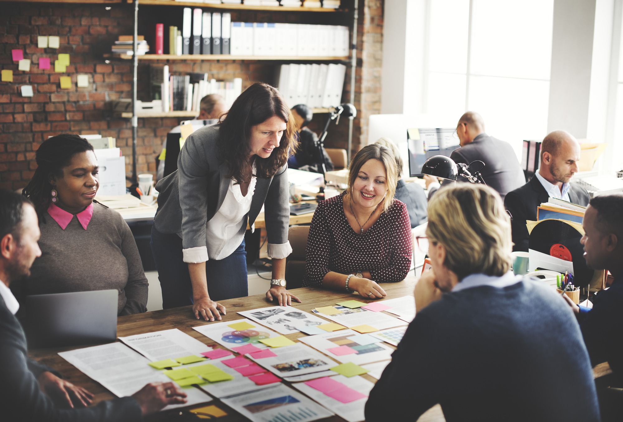 A diverse group collaborates around a table with documents and colorful sticky notes. The room has a bright, engaged atmosphere with shelves in the background.